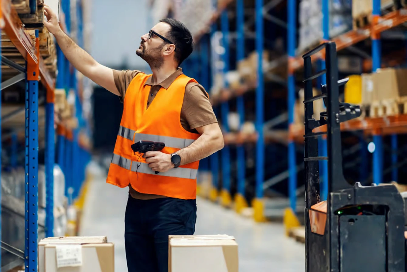 Logistics employee wearing safety vest using barcode scanner to manage stock in distribution center aisle