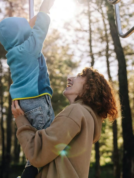 Mother lifting child toward playground bars outdoors with sunlight shining through forest trees